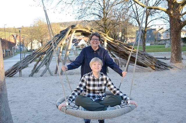 Eine Person sitzt auf einer Schaukel, eine zweite Person steht hinter ihr. Beide lachen und schauen Richtung Kamera.