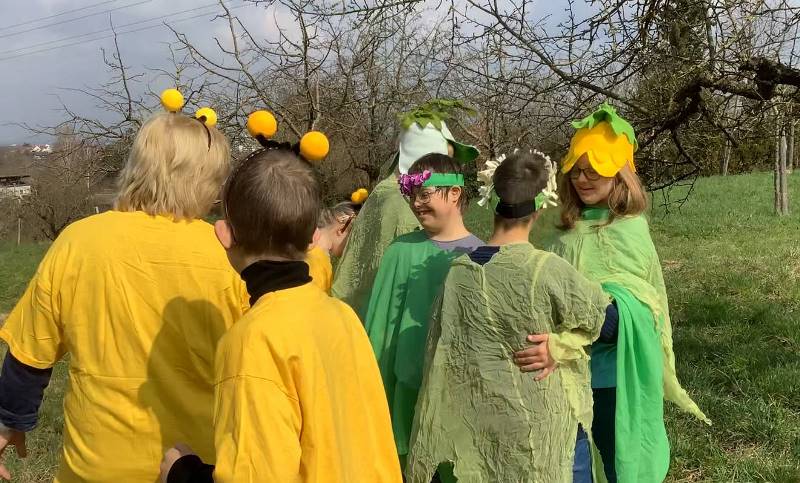 Eine Gruppe von Schülerinnen und Schüler der Theodor-Dierlamm-Schule steht in Blumen-, Bienen- und Schwalbenkostümen aus einem Nähprojekt auf einer Wiese mit Bäumen. 