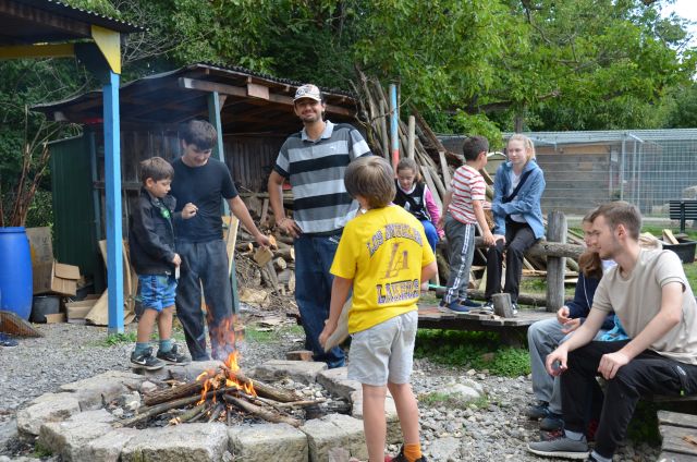 Mehrere Personen stehen und sitzen um ein Holzfeuer, das zwischen der runden Umgrenzung von großen Steinen am Boden brennt. Mehrere Personen stehen und sitzen um ein Holzfeuer, das zwischen der runden Umgrenzung von großen Steinen am Boden brennt.