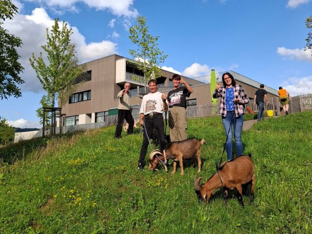 Vier Personen stehen auf einem mit Gras bewachsenen Hügel und winken Richtung Kamera. Zwei der Personen halten an Stricken insgesamt zwei braune Ziegen, die Gras fressen. Im Hintergrund sind weitere zwei Personen und Häuser zu sehen. 