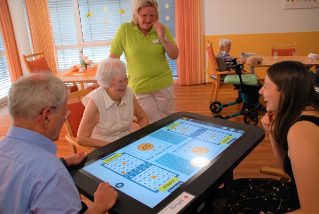 Drei Personen sitzen um ein CareTable und eine weitere Person steht dabei. Zwei der sitzenden Personen lachen sich gegenseitig an. Drei Personen sitzen um ein CareTable und eine weitere Person steht dabei. Zwei der sitzenden Personen lachen sich gegenseitig an.