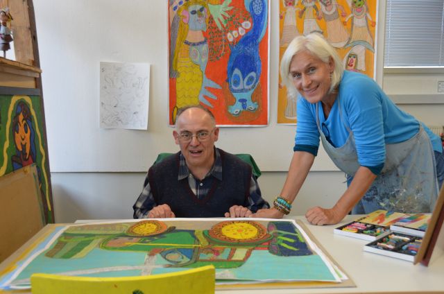 Eine Person sitzt an einem Tisch vor einem gemalten Bild, eine weitere Person steht neben ihr und beide lachen Richtung Kamera.