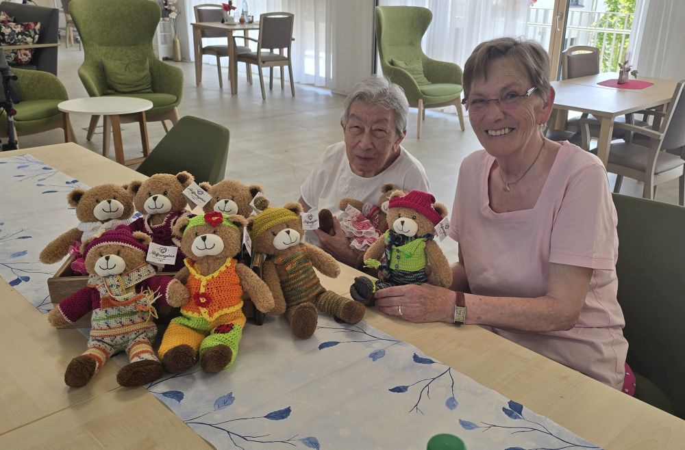 Zwei Personen sitzen an einem Tisch, auf dem eine Anzahl der von den Seniorinnen des Alexander-Stifts eingekleideten Hoffnungsbären zu sehen sind. Zwei Personen sitzen an einem Tisch, auf dem eine Anzahl der von den Seniorinnen des Alexander-Stifts eingekleideten Hoffnungsbären zu sehen sind.