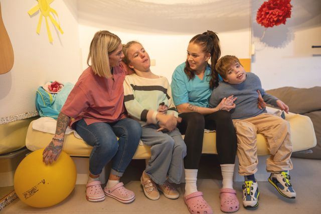 Zwei Heilerziehungspflegerinnen sitzen mit zwei Kindern der Kindergruppe KiWi auf einem Sofa und stützen die Kinder beim Sitzen. Zwei Heilerziehungspflegerinnen sitzen mit zwei Kindern der Kindergruppe KiWi auf einem Sofa und stützen die Kinder beim Sitzen.
