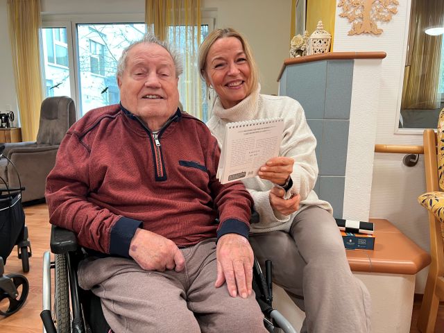 Zwei Personen sitzen nebeneinander und lachen Richtung Kamera. Eine der Personen sitzt im Rollstuhl.