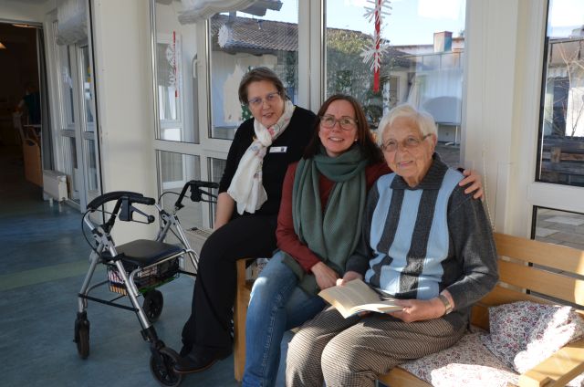 Drei Personen sitzen nebeneinander und blicken lächelnd Richtung Kamera. Neben ihnen steht ein Rollator.