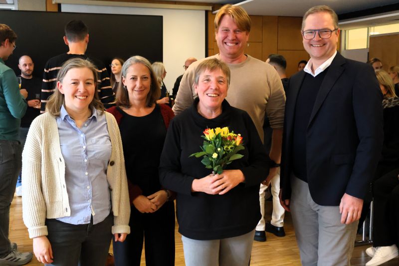 Fünf Personen stehen nebeneinander und lächeln Richtung Kamera. Die Person in der Mitte hält einen Blumenstrauß in ihren Händen.