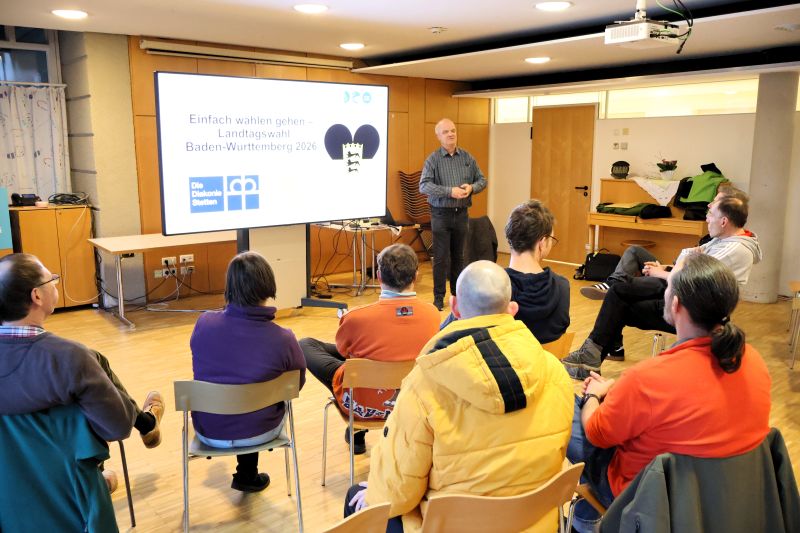 Der Referent der Info-Veranstaltung zur Wahl in Einfacher Sprache steht an einer Tafel, vor ihm sind einige der Teilnehmenden auf Stühlen sitzend zu sehen. Der Referent der Info-Veranstaltung zur Wahl in Einfacher Sprache steht an einer Tafel, vor ihm sind einige der Teilnehmenden auf Stühlen sitzend zu sehen.