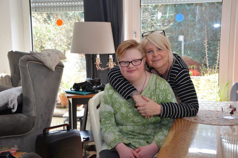 In einem Zimmer sitzt eine Person auf einem Stuhl. Eine zweite Person bückt sich von hinten zu ihr hinunter und legt ihre Arme um sie. 