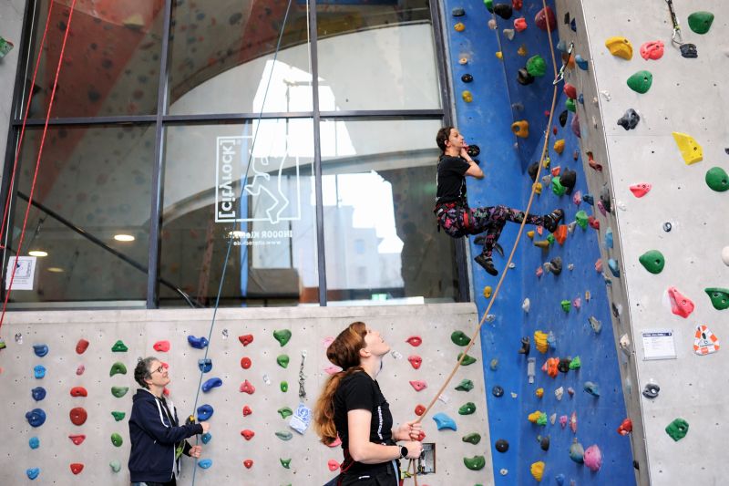 Eine junge Person klettert von zwei weiteren Personen gesichert in der Kletterhalle cityrock in Stuttgart Mitte. Im Hintergrund befindet sich eine Glaswand mit Logo und Aufschrift cityrock.