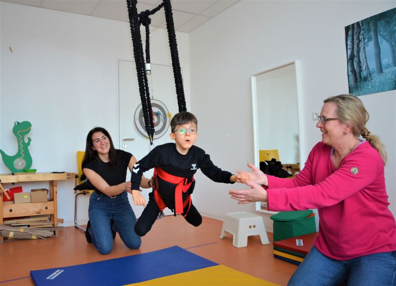Therapie-Situation in der Interdisziplinäre Frühförderstelle – Ein Kind schwingt in einem Geschirr in der Bungeeschaukel durch die Luft, zwei Personen unterstützen dabei.
