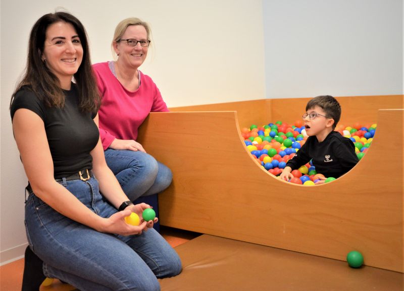 Therapie-Situation in der Interdisziplinäre Frühförderstelle – Ein Kind sitzt inmitten bunter Bälle, zwei Personen halten sich vor dem Bällebad auf.  