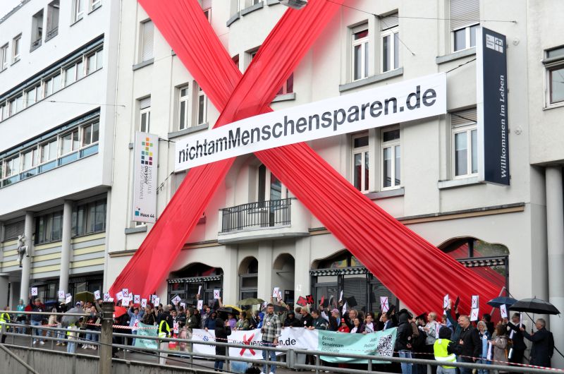 Protestaktion in Stuttgart am 5. Mai 2026. Zahlreiche Personen stehen vor einem Haus, das an der Front mit einem großen roten Kreuz bespannt ist und einem Banner mit der Aufschrift nichtamMenschensparen.de