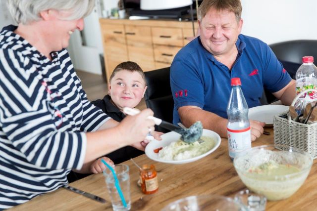 Ein Junge sitzt zwischen 2 erwachsen Personen an einem Tisch, eine der Personen schöpft Essen in einen Teller. 