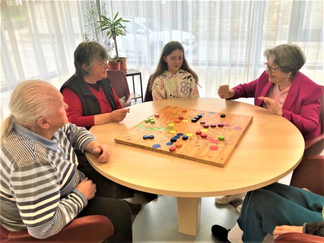 Mehrere Personen sitzen um einen Tisch, auf dem ein übergroßes „Mensch-ärgere-dich-nicht“-Spielbrett liegt. Mehrere Personen sitzen um einen Tisch, auf dem ein übergroßes „Mensch-ärgere-dich-nicht“-Spielbrett liegt.