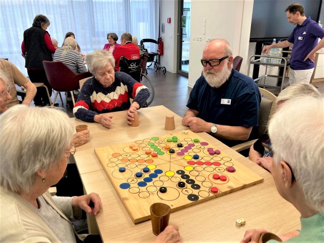 Mehrere Personen sitzen um einen Tisch, auf dem ein übergroßes „Mensch-ärgere-dich-nicht“-Spielbrett liegt. Mehrere Personen sitzen um einen Tisch, auf dem ein übergroßes „Mensch-ärgere-dich-nicht“-Spielbrett liegt.