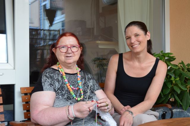 Zwei Personen sitzen nebeneinander und blicken Richtung Kamera, eine der Personen strickt. Zwei Personen sitzen nebeneinander und blicken Richtung Kamera, eine der Personen strickt.