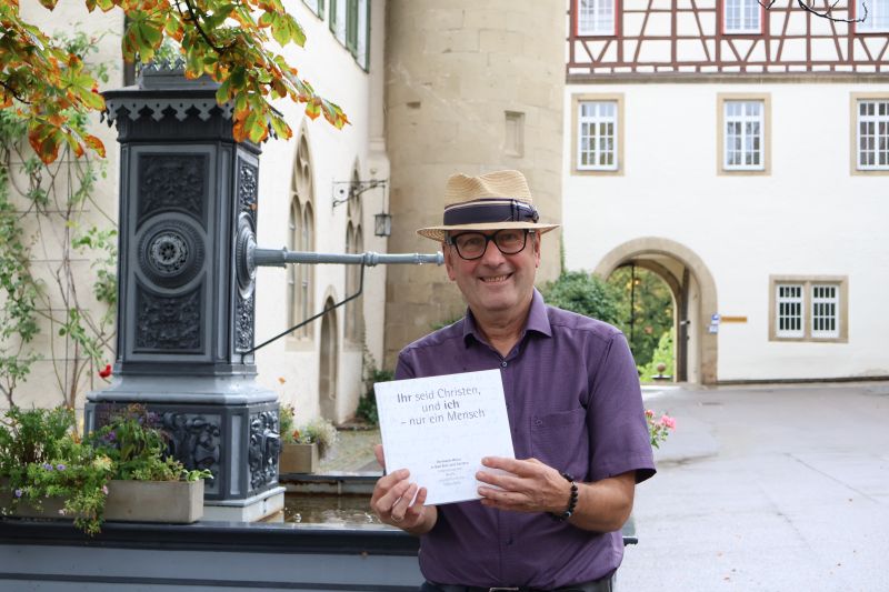 Eine Person steht vor dem Schlossbrunnen und hält die Broschüre „Ihr seid Christen, und ich – nur ein Mensch“ Richtung Kamera. Eine Person steht vor dem Schlossbrunnen und hält die Broschüre „Ihr seid Christen, und ich – nur ein Mensch“ Richtung Kamera.