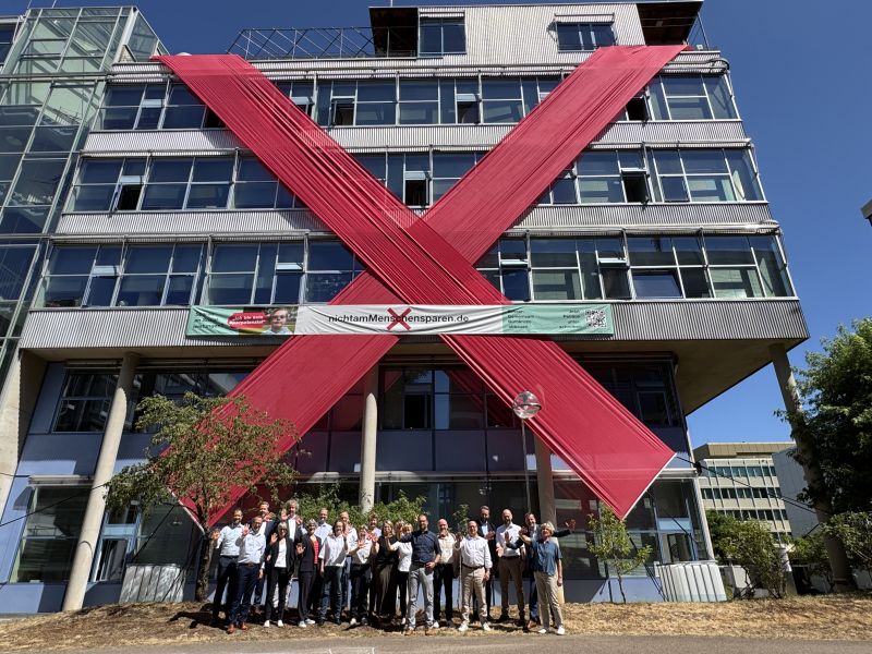 Ein großes rotes Stoff-Banner in X-Form ist an einem Gebäude angebracht. Quer darüber ist ein weiteres Banner angebracht mit der Aufschrift „Nicht am Menschen sparen.“ Unten am Gebäude steht eine Gruppe von Menschen. Ein großes rotes Stoff-Banner in X-Form ist an einem Gebäude angebracht. Quer darüber ist ein weiteres Banner angebracht mit der Aufschrift „Nicht am Menschen sparen.“ Unten am Gebäude steht eine Gruppe von Menschen.