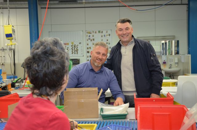 Zwei Personen an einem Arbeitstisch der Remstal Werkstätten am Standort Kernen-Stetten in der Mercedesstraße schauen Richtung Kamera. Eine dritte Person ist von hinten zu sehen. Zwei Personen an einem Arbeitstisch der Remstal Werkstätten am Standort Kernen-Stetten in der Mercedesstraße schauen Richtung Kamera. Eine dritte Person ist von hinten zu sehen.