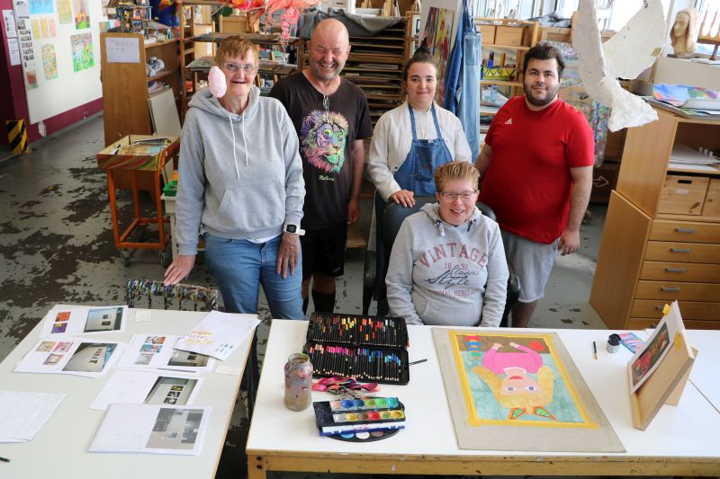 Fünf Künstler*innen und Künstler sitzen und stehen im Atelier der Kreativen Werkstatt. Fünf Künstler*innen und Künstler sitzen und stehen im Atelier der Kreativen Werkstatt.